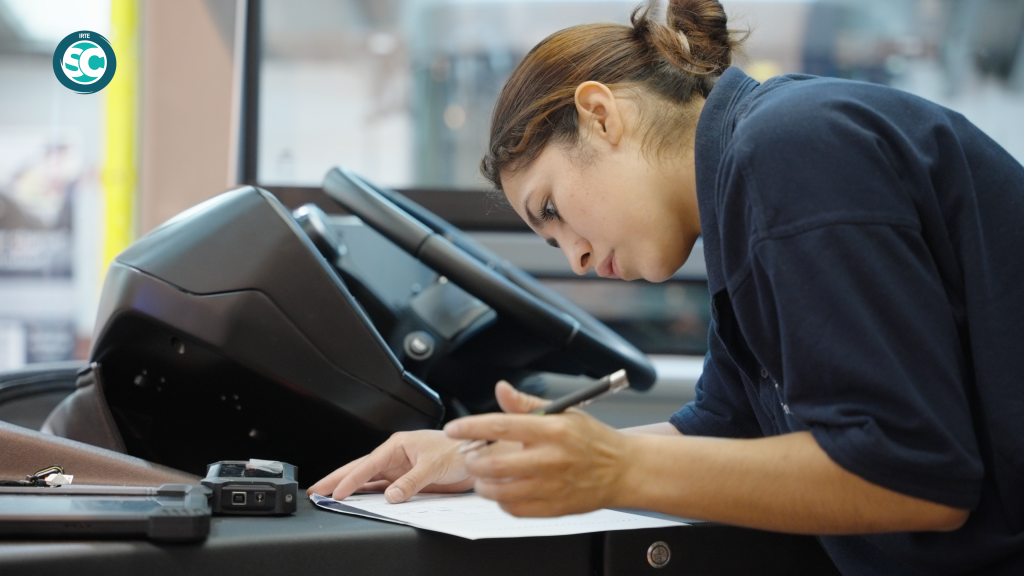 A person leaning over a desk, signing papers with a pen beside a payment terminal and card reader at the counter.