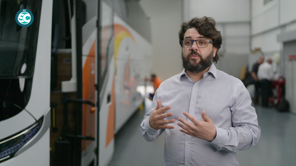 Bearded man with glasses in a light shirt gesturing while standing beside a large bus in a workshop environment.